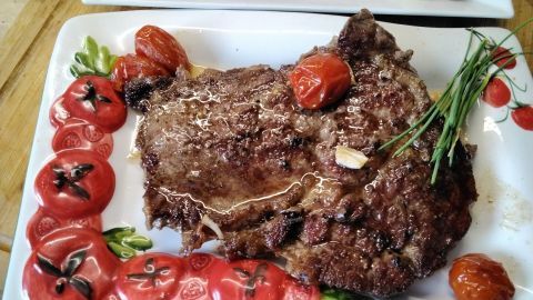 Ein saftiges Rib-Eye-Steak liegt auf einem dekorativen Teller mit roten Tomaten und Kräutern, umgeben von einer warmen Küche.