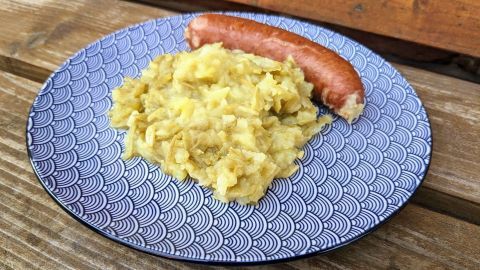 Ein Teller mit Fassbohnen-Eintopf zeigt eine Portion aus zerdrückten Bohnen und eine Wurst, auf einer Holzoberfläche.