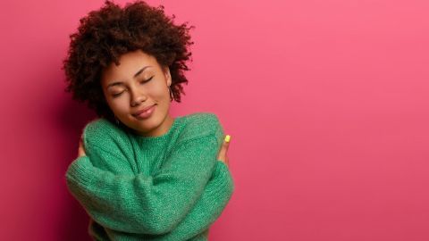 Eine junge Frau mit lockigem Haar umarmt sich selbst in einem grünen Pullover vor einem rosa Hintergrund, was Wohlbefinden strahlt.