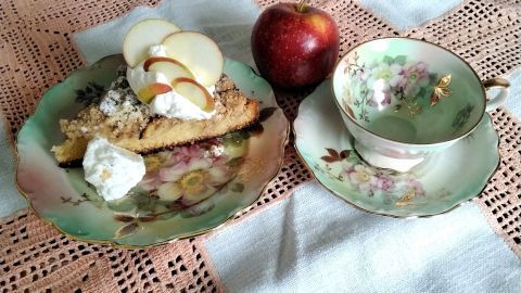 Ein Stück Apfelkuchen mit Zimt-Streuseln und Sahne auf einem dekorativen Teller neben einem roten Apfel und einer Tasse.