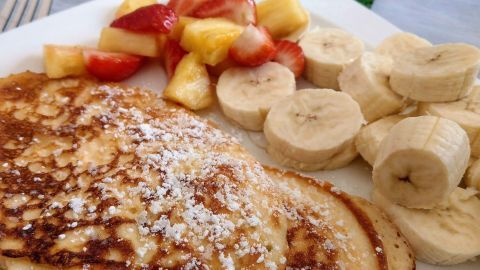 Auf einem weißen Teller liegen goldbraune, proteinreiche Pfannkuchen, umgeben von frischen Früchten wie Banane und Erdbeere.