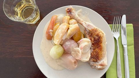 Ein gebratenes Huhn liegt auf einem Teller, umgeben von buntem Gemüse in einer cremigen Sauce, auf einem Holztisch.
