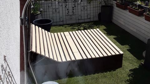 Eine Gartenliege mit Lattenrost steht in einem gepflegten Garten, umgeben von Pflanzen und bei sonnigem Wetter.