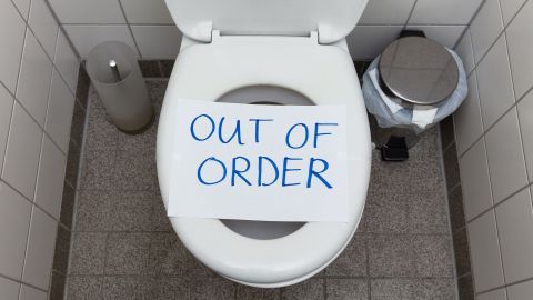 Eine Toillette mit einem Schild "OUT OF ORDER" darauf, steht in einem modernen Badezimmer mit einer offenen Abfalltonne nebenan.