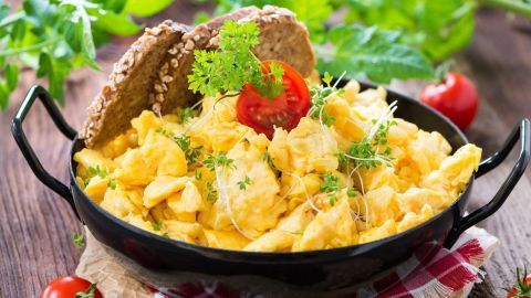 Rührei wird in einer schwarzen Pfanne serviert, garniert mit Tomate und Kräutern, neben zwei Brotscheiben.