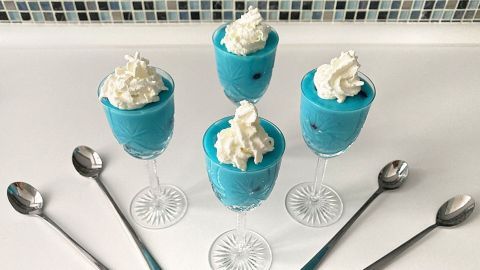 Blauäugiger Schlumpf-Pudding in eleganten Gläsern mit Sahnehaube steht vor einem Mosaik-Hintergrund auf einer weißen Fläche.