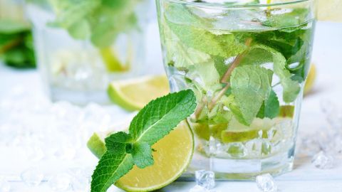 Ein Glas erfrischendes Getränk mit Pfefferminze und Limette steht auf einem hellen Tisch, umgeben von Eiswürfeln.