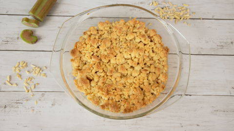 Ein goldbrauner Rhabarber-Crumble in einer Glasform steht auf einem Holzschirm, umgeben von Rhabarberstücken und Mandelsplittern.