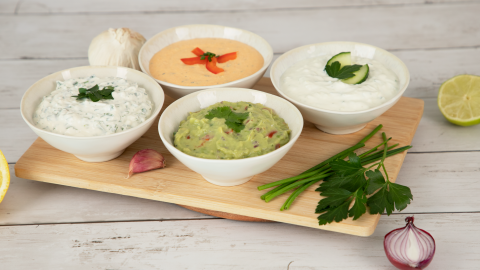 Vier Schalen mit verschiedenen Dips zum Grillen stehen auf einem Holzbrett, umgeben von frischen Zutaten wie Knoblauch, Zwiebel und Zitrusfrüchten.