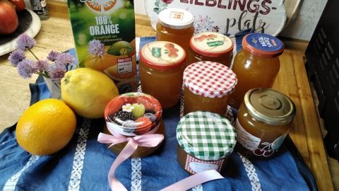 Gläser mit Holunderblüten-Orangengelee und frischen Früchten liegen auf einem blauen Tuch, umgeben von einer Obstschale.