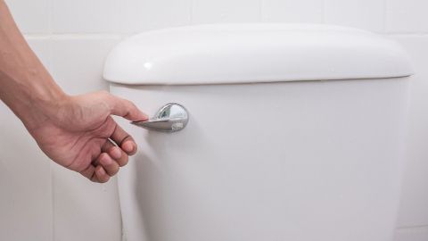 Eine Hand betätigt den Spülknopf einer weißen Toilette in einem hellen Bad, um Wasser für die Reinigung zu aktivieren.