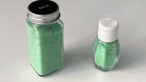 Zwei Gläser mit grünem Salz stehen nebeneinander auf einem hellen Untergrund, bereit zum Würzen von Speisen.