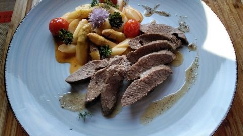 Zarte Lammlachs-Scheiben liegen dekorativ auf einem Teller mit Spargelragout und bunten Tomaten in einem feinen Sud.