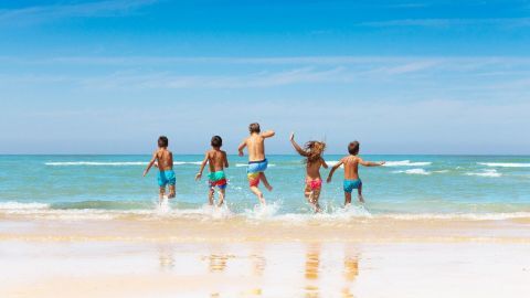 Fünf Kinder laufen fröhlich ins Meer und spritzen Wasser, während sie am sonnigen Strand spielen.