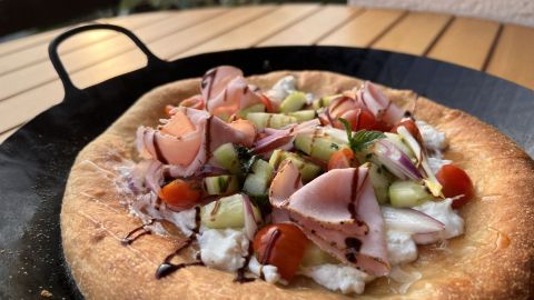 Eine knusprige Pizza ist mit frischem Salat, Schinken, Gemüse und einer Balsamico-Reduktion belegt, serviert auf einem Tisch.