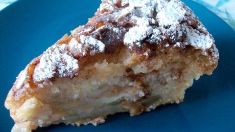 Ein Stück Apfel-Rührkuchen liegt auf einem blauen Teller, leicht mit Puderzucker bestäubt, im Hintergrund ein grünes Tuch.