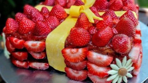 Ein wunderschöner Erdbeerkuchen in Geschenkform, dekoriert mit frischen Erdbeeren und einer gelben Schleife, umgeben von Natur.