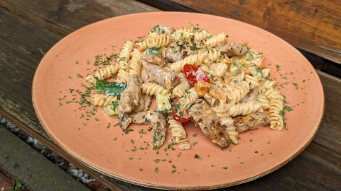 Ein Teller Gyros-Nudelsalat zeigt gefüllte Pasta und Fleischstücke mit frischem Gemüse, angerichtet auf einem rustikalen Holzuntergrund.