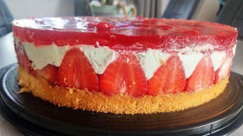 Eine bunte Erdbeer-Waldmeistertorte steht auf einem schwarzen Teller, umgeben von einem hellen, modernen Raum.