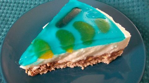 Ein Stück No-Bake Cheesecake mit Quark steht auf einem Teller, umgeben von einer bunten, gelierten Schicht.