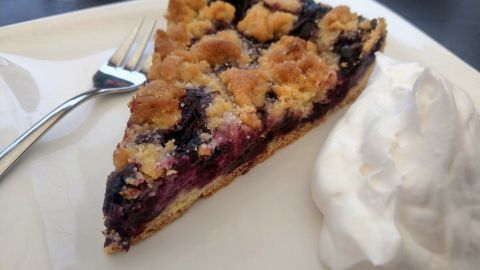 Ein Stück Heidelbeerkuchen mit Streuseln liegt auf einem Teller, daneben steht ein Klecks Schlagsahne.