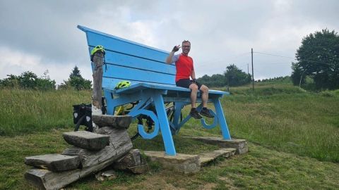 Eine große, blaue Bank steht auf einer Wiese, während ein Mann in rotem Shirt lächelnd und winkend darauf sitzt.
