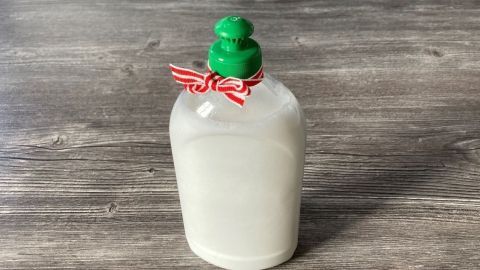 Ein flacher Behälter mit grünem Verschluss und rotem Band steht auf einer Holzoberfläche, gefüllt mit selbstgemachtem Spülmittel.
