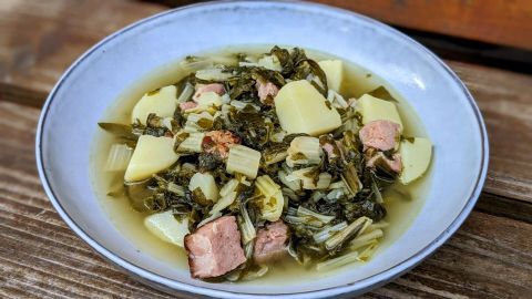 Eine Schüssel Stielmus-Suppe liegt auf einem Holzuntergrund, gefüllt mit Gemüse, Kartoffeln und Fleischstücken.