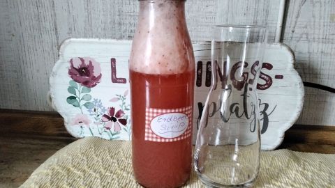 Eine Flasche mit selbstgemachtem Erdbeersirup steht neben einem Glas auf einem holzverkleideten Tisch, dekoriert mit Blumen.