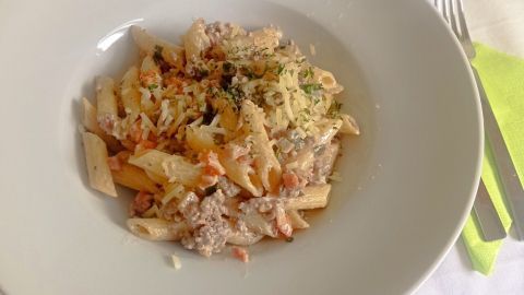 Ein Teller Penne mit Ragú Bianco, garniert mit Käse und Kräutern, auf einem Tisch mit Besteck und Serviette.
