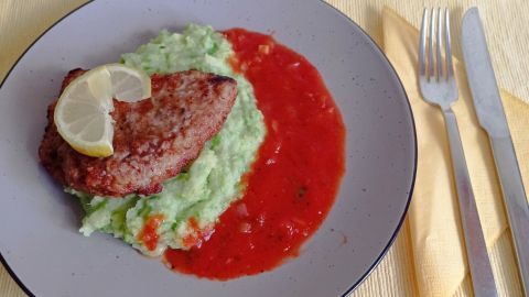 Ein goldbraun gebratenes Wiener Schnitzel liegt auf grünem Kartoffel-Erbsen-Püree, umgeben von roter Tomatensoße.