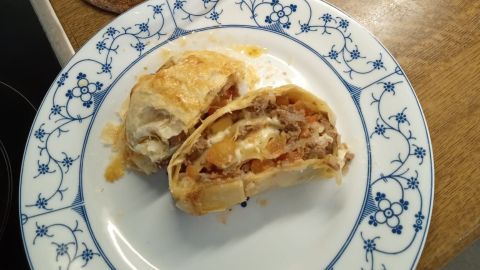 Blätterteigrollen sind goldbraun gebacken und gefüllt mit Fleisch und Gemüse auf einem Teller neben einer Auflaufform.