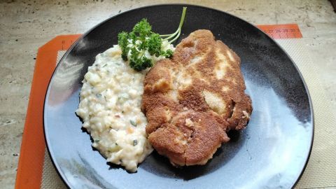 Ein gebratenes Schweinekamm-Schnitzel liegt neben cremigem Kartoffelsalat auf einem schwarzen Teller.