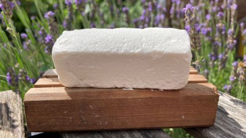 Ein Stück festes Spülmittel liegt auf einem Holzbrett, umgeben von lila Lavendelpflanzen im Hintergrund.