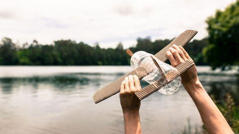 Ein Kind hält ein selbstgebautes Flugzeug über einem ruhigen Gewässer, bereit zum Abheben in einer grünen Umgebung.