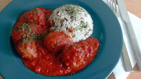 Saftige Hackbällchen in Tomatensauce sind auf einem blauen Teller, serviert mit Reis und frisch gehackten Kräutern.
