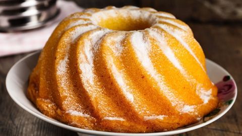 Ein goldbrauner Zitronen-Rührkuchen als Gugelhupf steht auf einem weißen Teller mit blumenförmiger Verzierung.