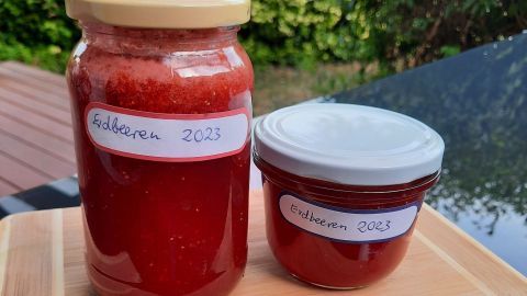 Ein großes und ein kleines Glas mit selbstgemachter Erdbeermarmelade stehen auf einem Holzbrett in einem grünen Garten.