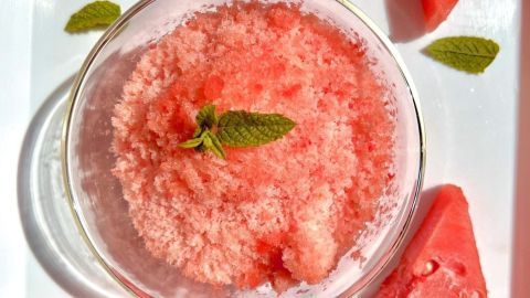 Eine Schüssel mit frischem, rosa Shaved Ice aus Wassermelone ist von Wassertropfen und Melonenstücken umgeben.