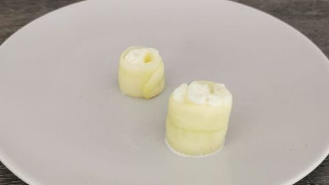 Auf einem weißen Teller liegen zwei Sushi-Rollen aus Gurke, gefüllt mit einer cremigen Füllung, auf einem Holzuntergrund.