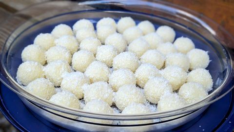 Runde, weiße Raffaello-Kugeln stehen in einer Glasschüssel, umgeben von flauschiger Kokosraspel, auf einem Holzuntergrund.
