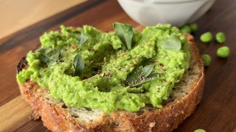 Ein Stück Vollkornbrot ist mit leuchtend grüner Erbsen-Guacamole bestrichen und von grünen Erbsen umgeben.