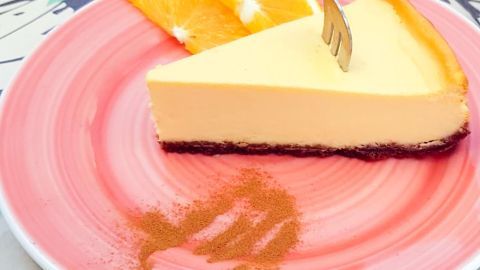 Ein Stück amerikanischer Käsekuchen auf einem rosa Teller. Einsteckende Gabel, verziert mit Orangenscheiben und Zimt.