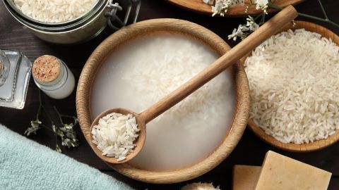 Ein Holzlöffel hebt Reiswasser aus einer Schale, umgeben von Reis, einem Handtuch und Naturprodukten für die Haarpflege.