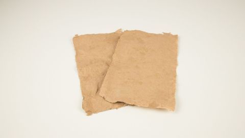 Zwei selbst geschöpfte Papierteile liegen auf einer weißen Oberfläche und zeigen eine unregelmäßige, natürliche Textur.