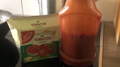 Eine Flasche mit Tomatensoße steht neben einer Packung passierten Tomaten auf einem Herd in einer modernen Küche.