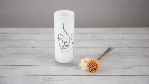 Eine weiße Vase mit einer schwarzen Linienzeichnung eines Gesichts steht auf einem Holztablett, daneben ein getrocknetes Blumenarrangement.