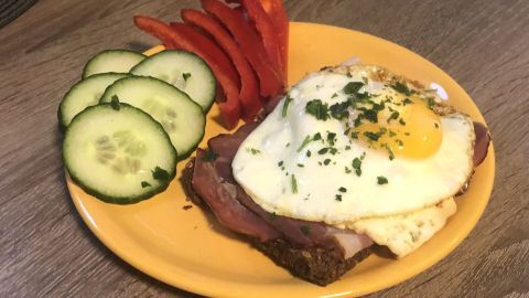 Ein belegtes Brot mit Schwarzwälder Schinken, Spiegelei und frischem Gemüse in einem gelben Teller auf Holzoberfläche.