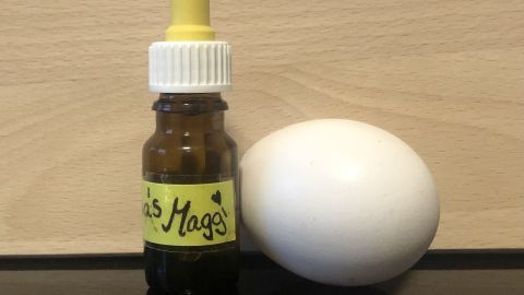Eine kleine Flasche mit gelbem Deckel steht neben einem weißen Ei auf einem Tisch aus hellem Holz, bereit zum Mitnehmen.