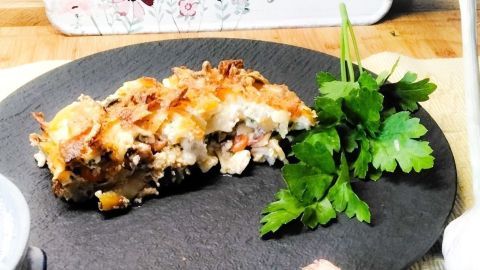 Ein Nudelauflauf mit Gemüse und Steinpilzen liegt auf einem schwarzen Teller, umgeben von frischen Kräutern und Knoblauch.